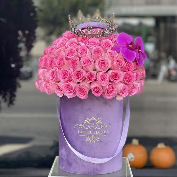 Pink roses in a purple hatbox with a jeweled crown and orchid accent