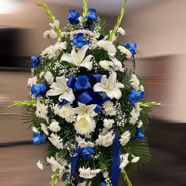 Tall standing spray of blue roses, white lilies, and white mums with blue ribbon on an easel.