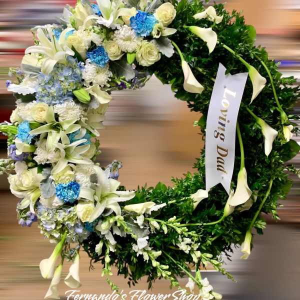 Round sympathy wreath with white lilies, roses, blue flowers, and a Loving Dad ribbon.