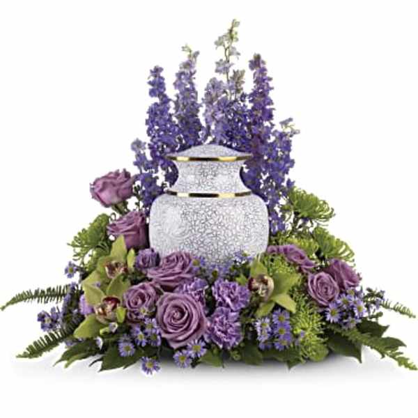 Lavender and purple floral arrangement surrounding a white cremation urn with tall delphinium accents.