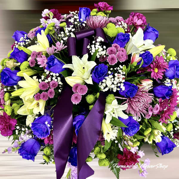 Large oval arrangement of purple, blue, white, and pink flowers with a purple ribbon bow
