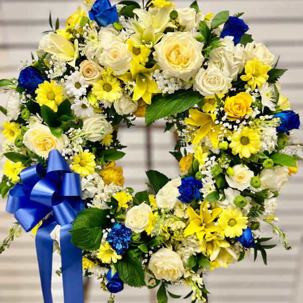 Standing wreath of white, yellow, and blue flowers with a large blue bow on an easel.