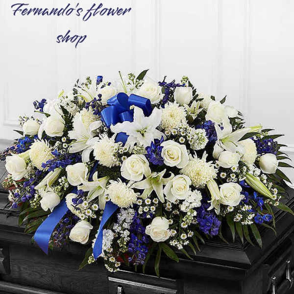 White and blue funeral casket spray with roses, lilies, and ribbon on a dark wood casket