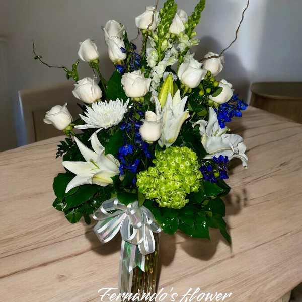 Tall arrangement of white roses and lilies with blue accents in a glass vase with a ribbon bow.