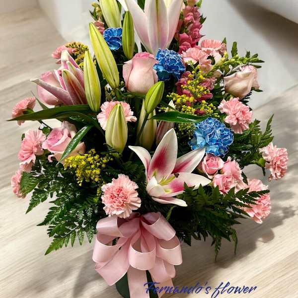 Tall arrangement of pink lilies, roses, carnations, and blue flowers with a pink bow in a green vase