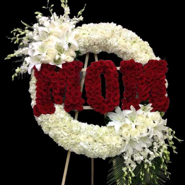 Large white standing wreath with red rose letters spelling MOM and white lilies on an easel