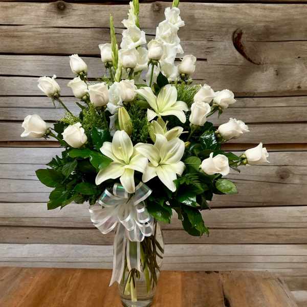 Tall clear vase of white roses, lilies, and snapdragons with a white ribbon bow.