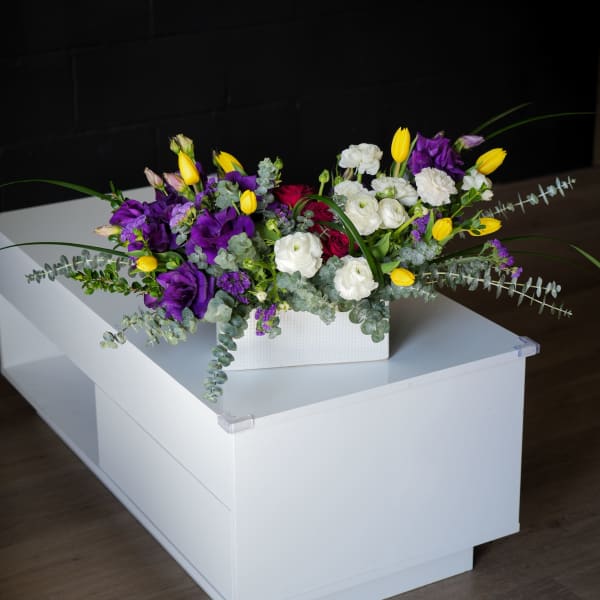 Low floral arrangement in a white rectangular box with purple, white, yellow, and red blooms