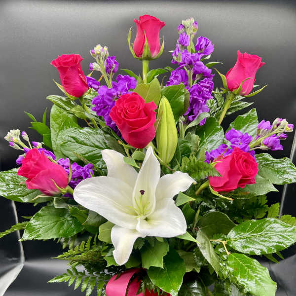 Arrangement of hot pink roses, purple stock, and white lilies in a purple vase with a pink bow