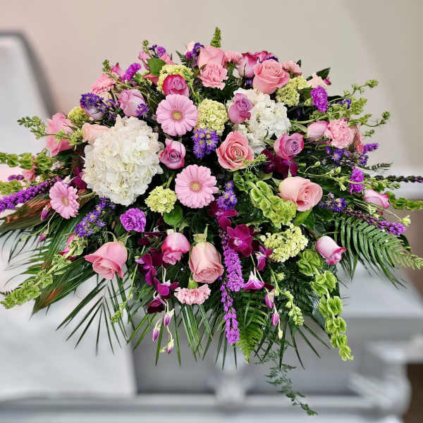 Pink and purple casket spray with roses, gerbera daisies, orchids, and hydrangeas on a white casket.
