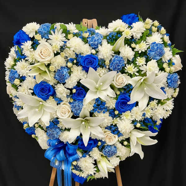 Heart-shaped blue and white standing floral wreath with roses, lilies, and carnations on a wooden easel.