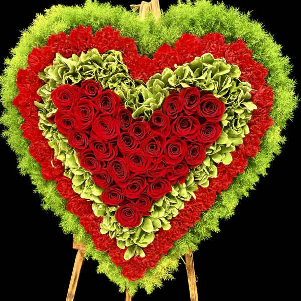 Heart-shaped standing floral spray of red roses with green and red flower borders on a wooden easel.