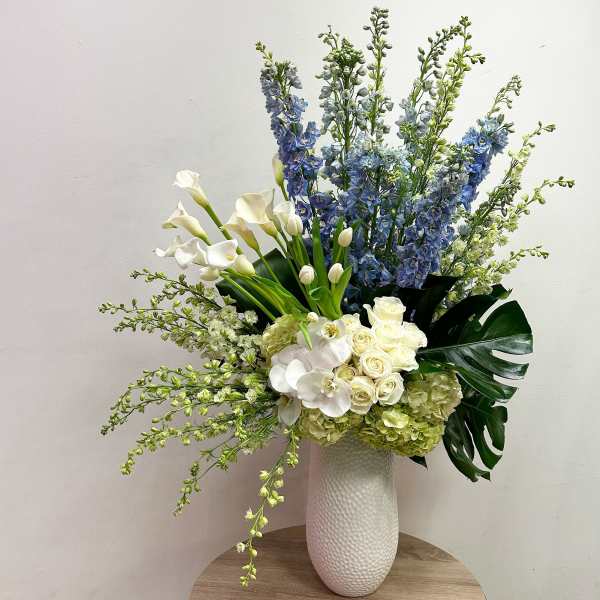 Tall arrangement of white roses, orchids, calla lilies and blue delphinium in a white vase
