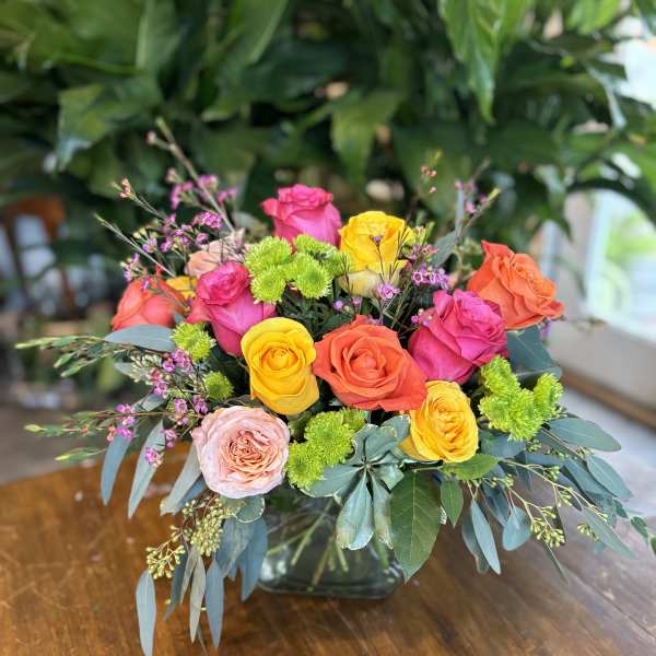 Bright arrangement of pink, orange and yellow roses with green accents in a clear glass cube vase.
