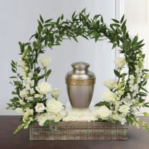 White roses and greenery arranged around a metal urn in a wooden box