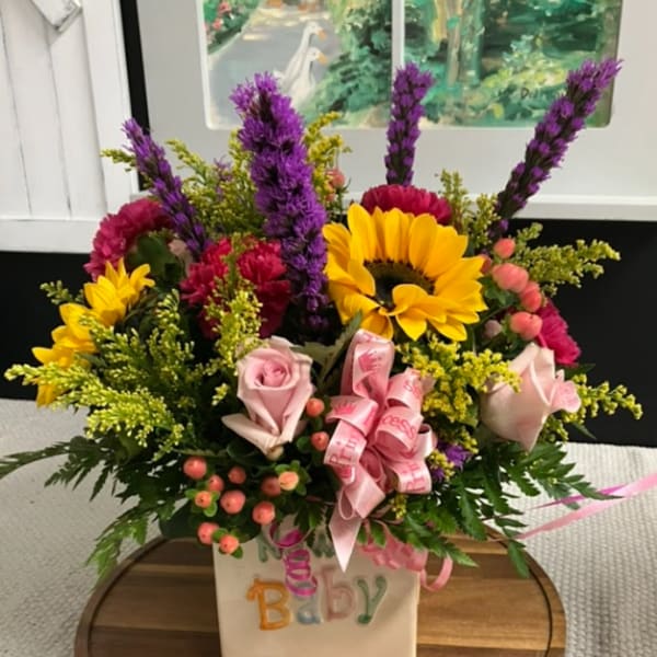Colorful baby-themed flower arrangement in a white vase with pink ribbon