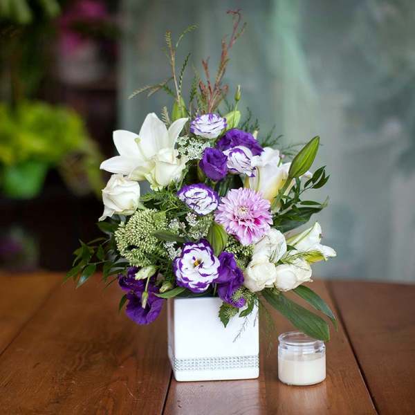 Purple and white floral arrangement in a white square vase beside a small candle