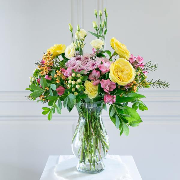 Mixed bouquet of yellow and pink flowers in a clear glass vase on a white surface