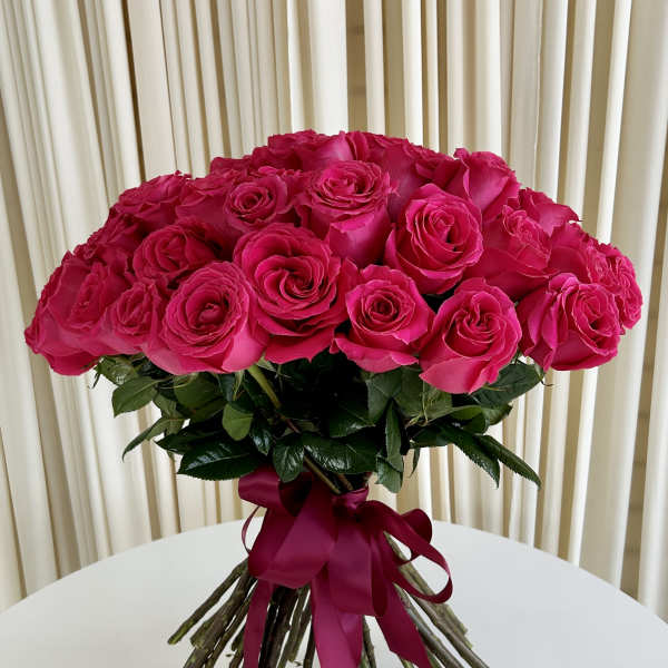 Hand-tied bouquet of bright pink roses with exposed stems and a matching ribbon on a white table.