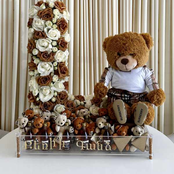 Tall number-shaped arrangement of white and caramel roses with teddy bears and mini bottles on a clear tray.