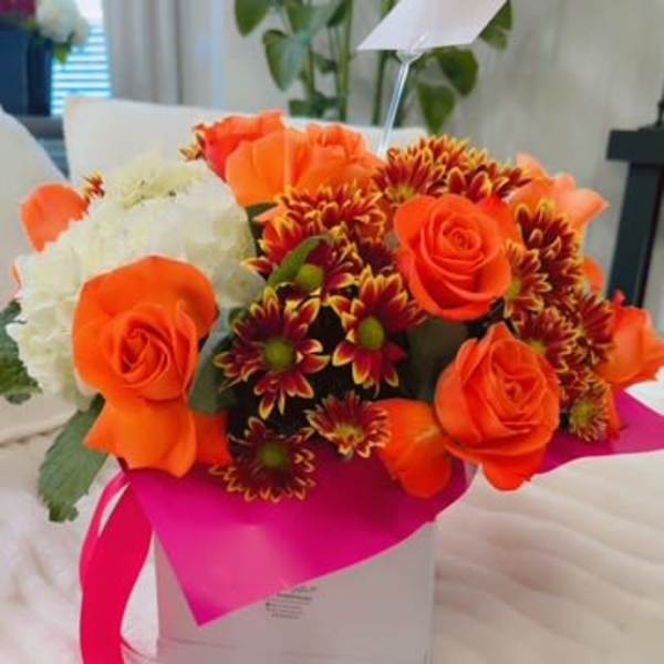 Orange roses and burgundy daisies in a white gift box with a pink ribbon.