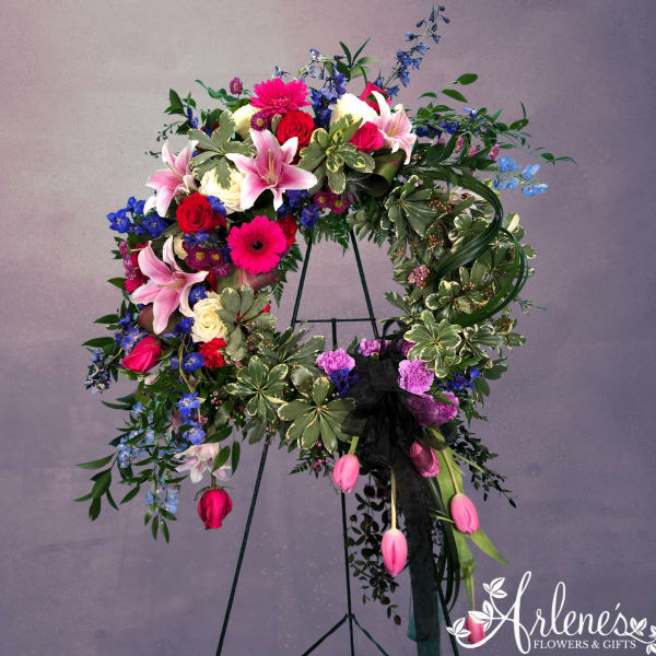 Standing wreath of pink lilies, roses, gerbera daisies and bright blooms on a ring of variegated foliage.