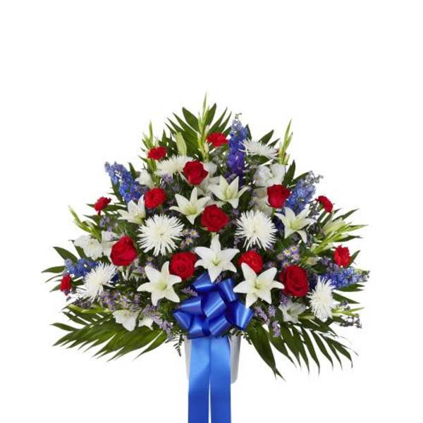 Fan-shaped red, white, and blue arrangement with roses, lilies, mums, and a blue ribbon in a white urn.