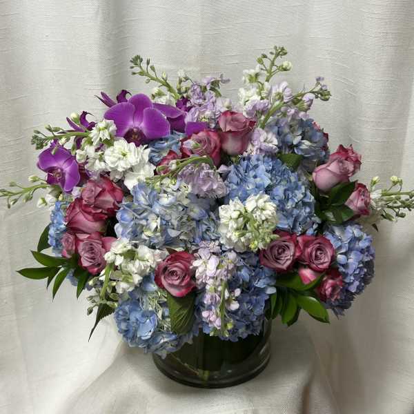 Lush arrangement of blue hydrangeas, pink roses, white stock, and purple orchids in a clear glass vase