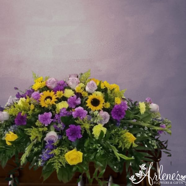 Colorful casket spray of yellow sunflowers, purple mums, and lavender and yellow roses on a wooden casket.