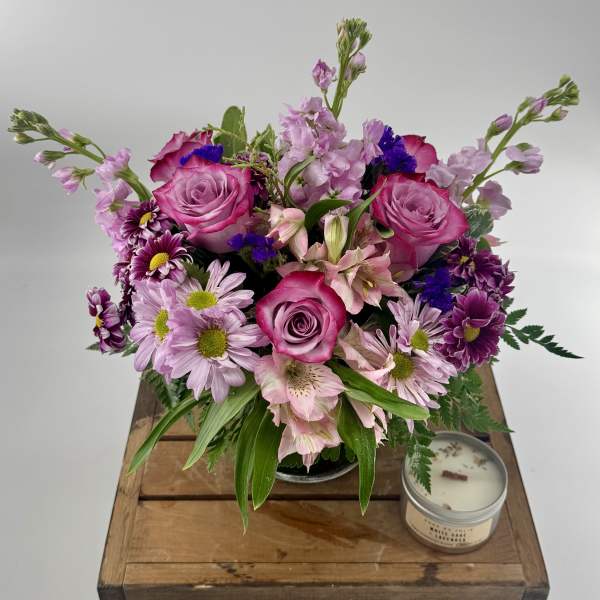 Round bouquet of pink roses, lavender mums, and alstroemeria in a glass vase beside a small candle.