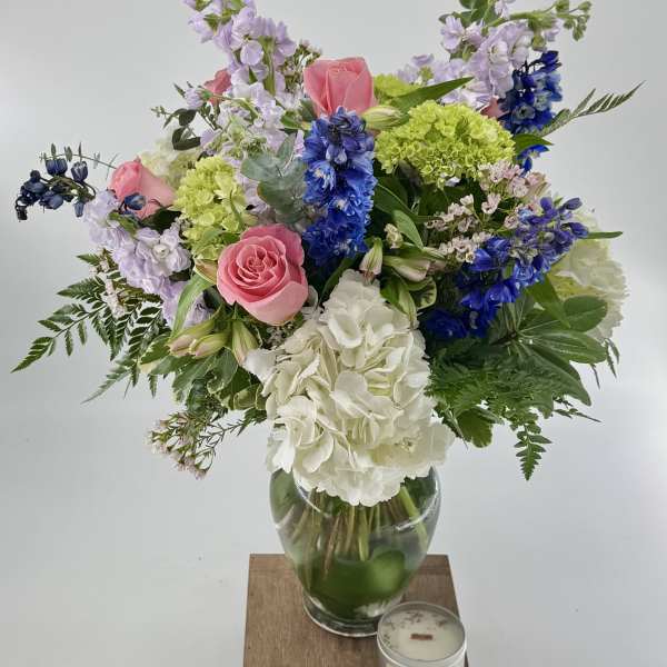Mixed bouquet of pink roses, blue delphinium, and white hydrangeas in a glass vase with a small candle.