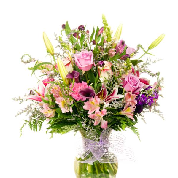 Tall mixed bouquet of pink roses, lilies, and purple accents in a clear glass vase with a mesh bow.