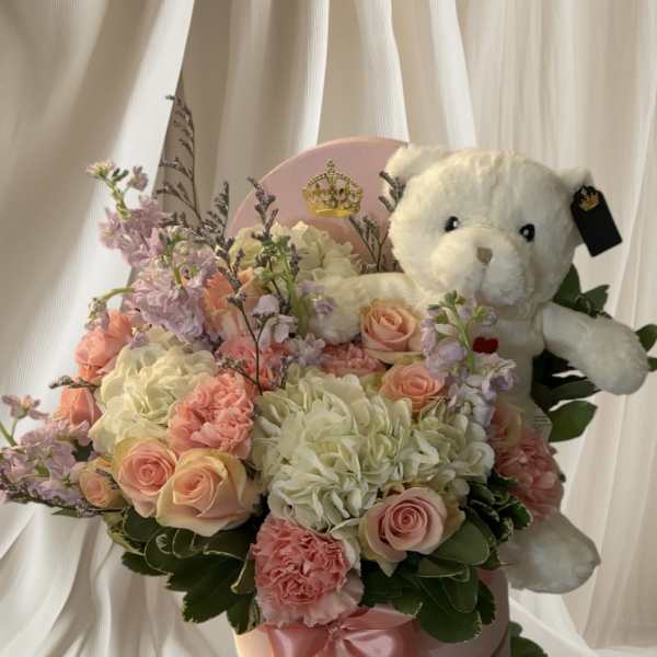 Pink and cream flower arrangement with a teddy bear in a round box