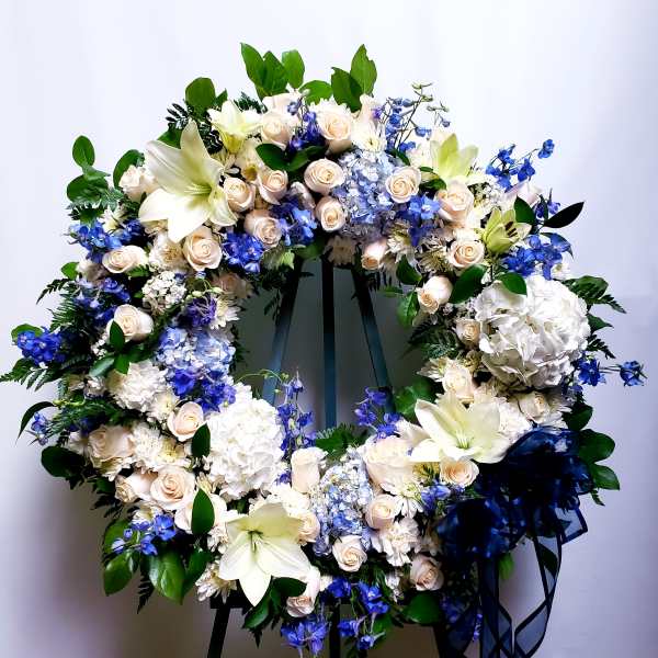 Large floral wreath with white lilies, cream roses, blue flowers, and a navy ribbon