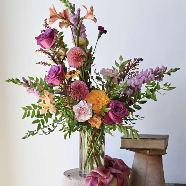 Tall mixed arrangement of purple, pink, and orange flowers in a clear glass vase beside stacked books.