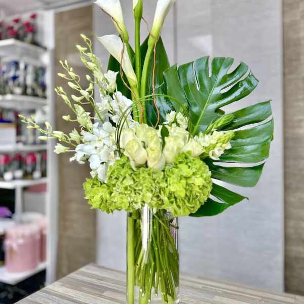 Tall white and green arrangement with calla lilies, orchids, and hydrangeas in a clear glass cylinder vase