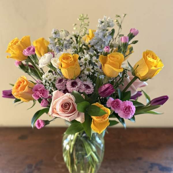 Mixed bouquet of yellow roses, pink roses, purple tulips, and small white daisies in a clear glass vase