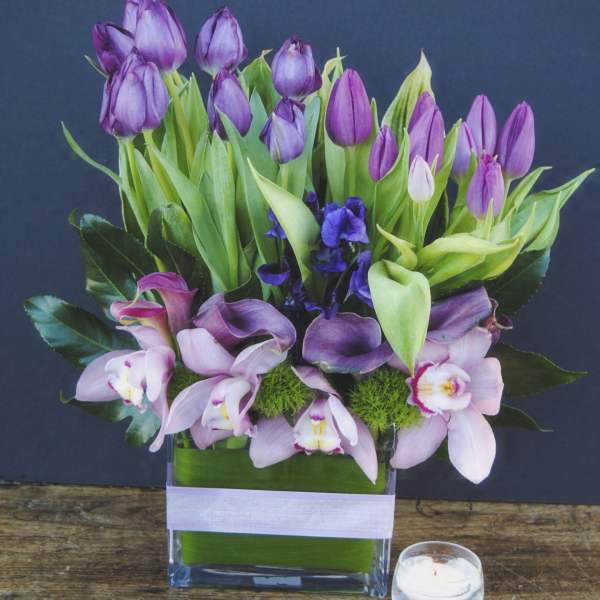 Purple tulips and pink orchids arranged in a glass vase with a small candle nearby