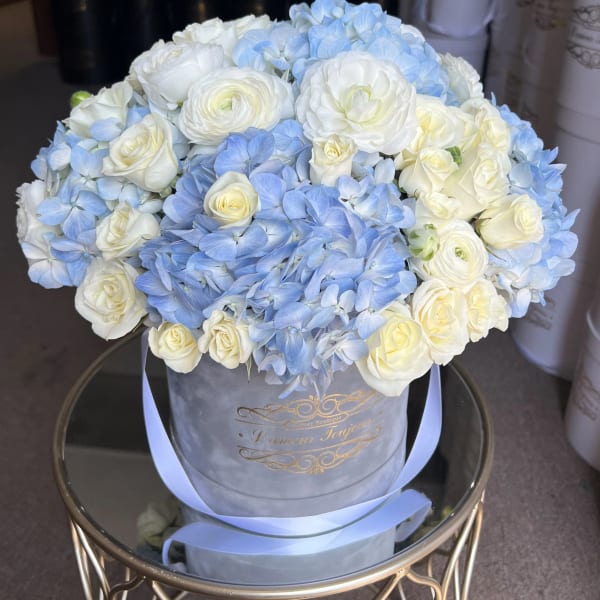 Round hatbox arrangement of white roses and ranunculus with light blue hydrangeas on a mirrored stand