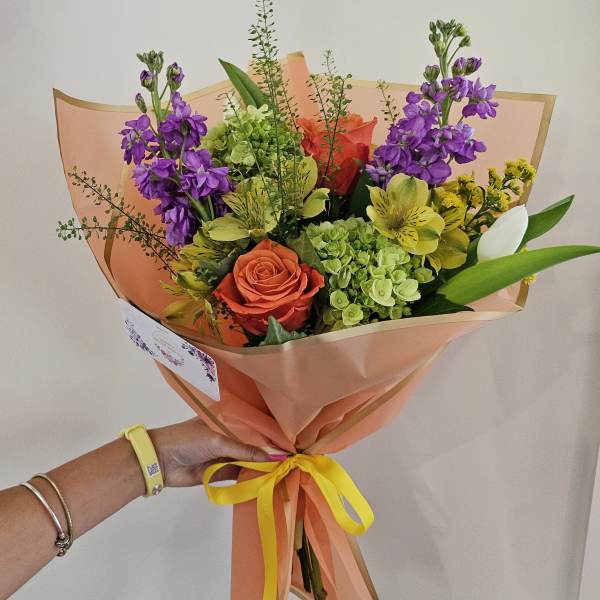 Handheld bouquet of purple, orange, yellow, and green flowers wrapped in peach paper