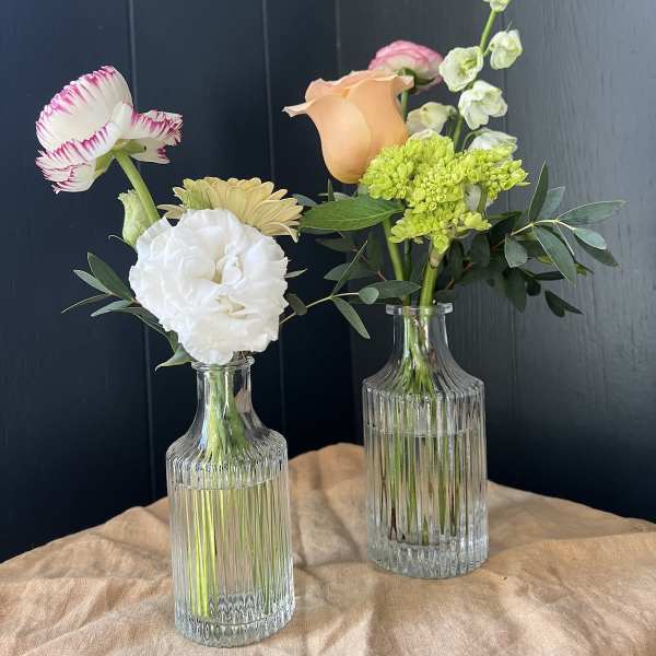 Two small floral arrangements in clear glass bottles with mixed blooms.