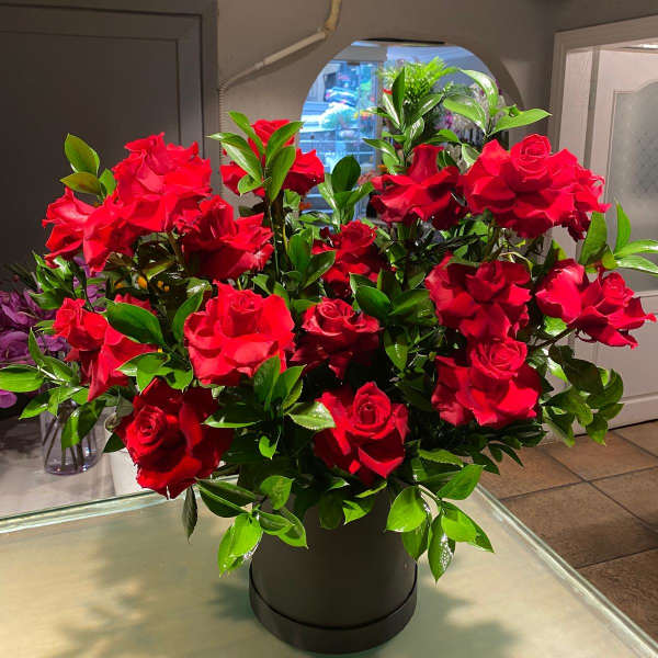 Red roses arranged in a black hatbox with glossy green foliage.
