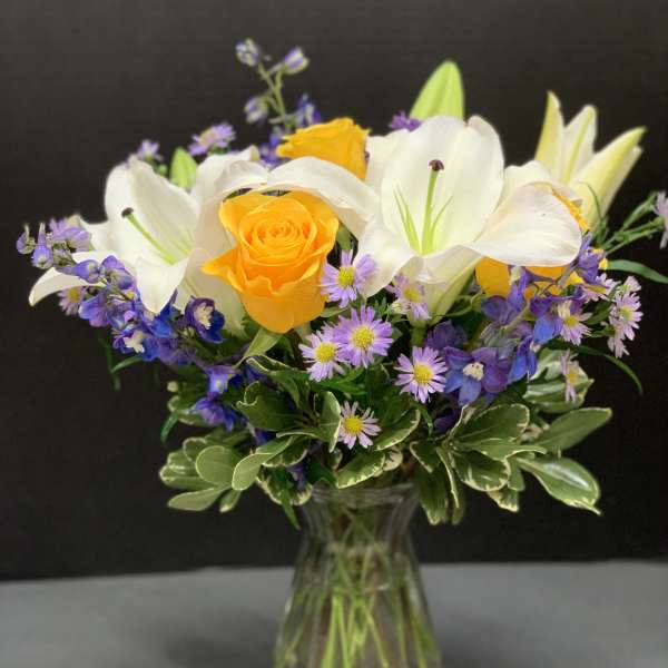 Bouquet of yellow roses, white lilies, and purple flowers in a glass vase