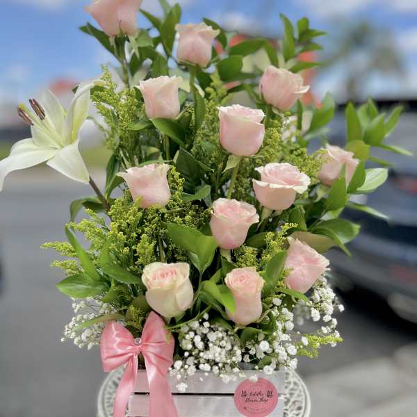 Pink roses and a white lily arranged in a wooden box with a pink ribbon.