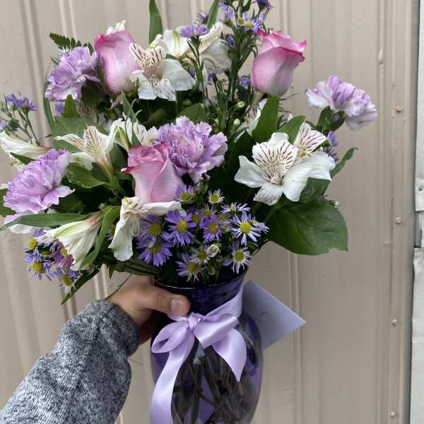 Handheld arrangement of pink roses and purple flowers in a purple glass vase with ribbon.
