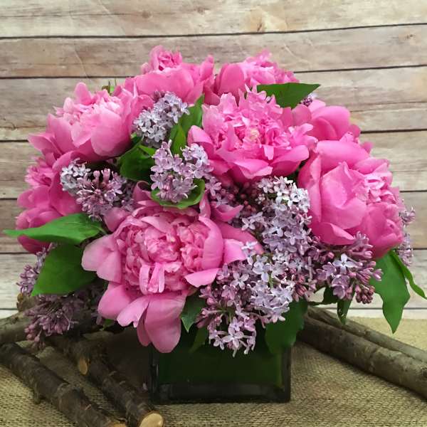 Pink peonies and lilac flowers arranged in a square glass vase