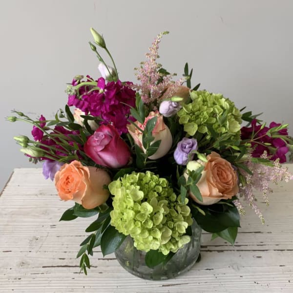 Low mixed arrangement of peach and pink roses, green hydrangeas, and purple blooms in a clear round vase