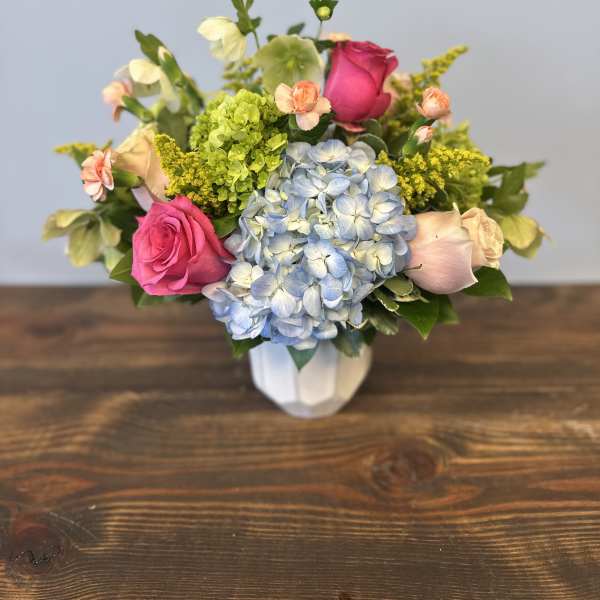 Compact arrangement of pink roses, blue hydrangeas, and mixed blooms in a white vase.