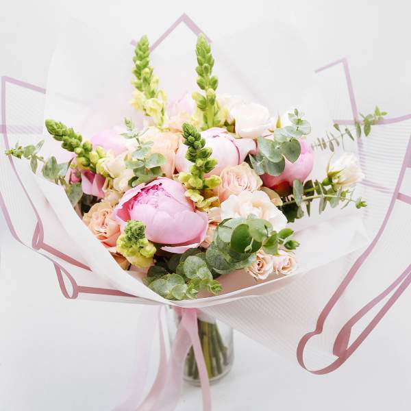 Hand-tied bouquet of pink peonies, peach roses, and snapdragons wrapped in white paper in a glass vase