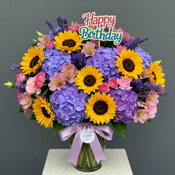 Bouquet of sunflowers, purple hydrangeas, and pink flowers in a glass vase with a birthday sign.